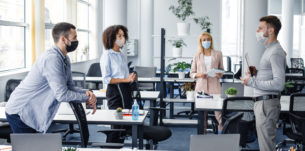 Modern team meeting, group work and social distancing. Manager with tablet speaks with workers in protective masks in interior of modern office with gadgets during coronavirus epidemic, free space